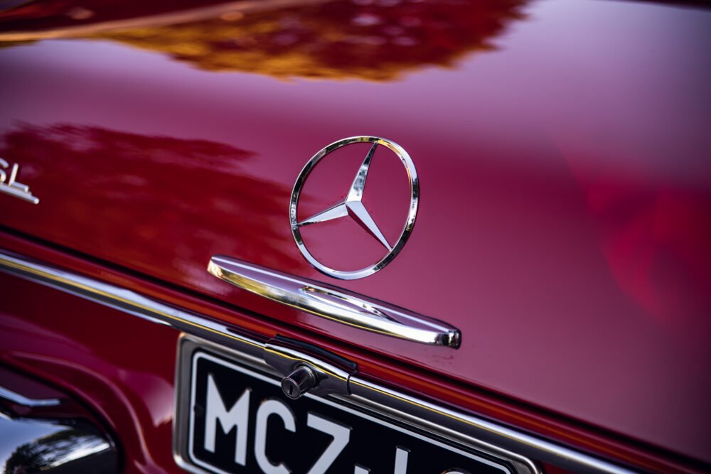 Close-up of vintage Mercedes hood emblem on red car.