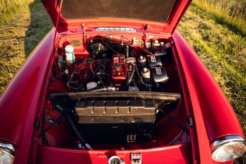 Red classic car engine compartment open in field.