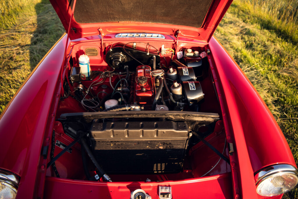 Red classic car engine compartment open in field.