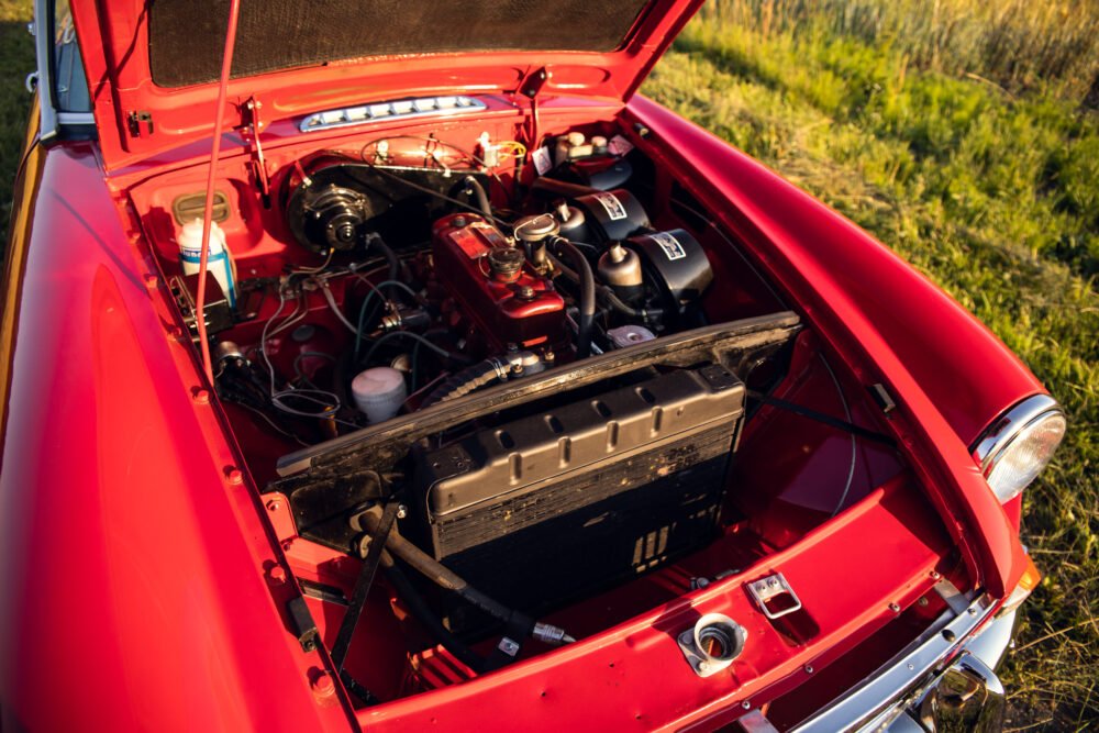 Red classic car's open hood showing engine in field.