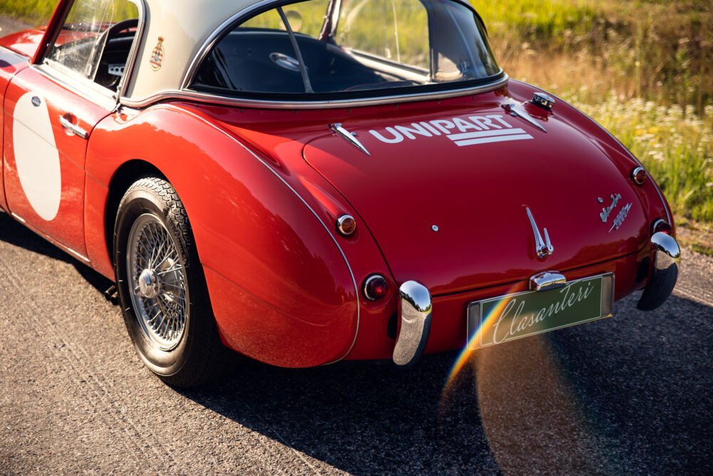 Red vintage car with racing decals, sunny roadside.