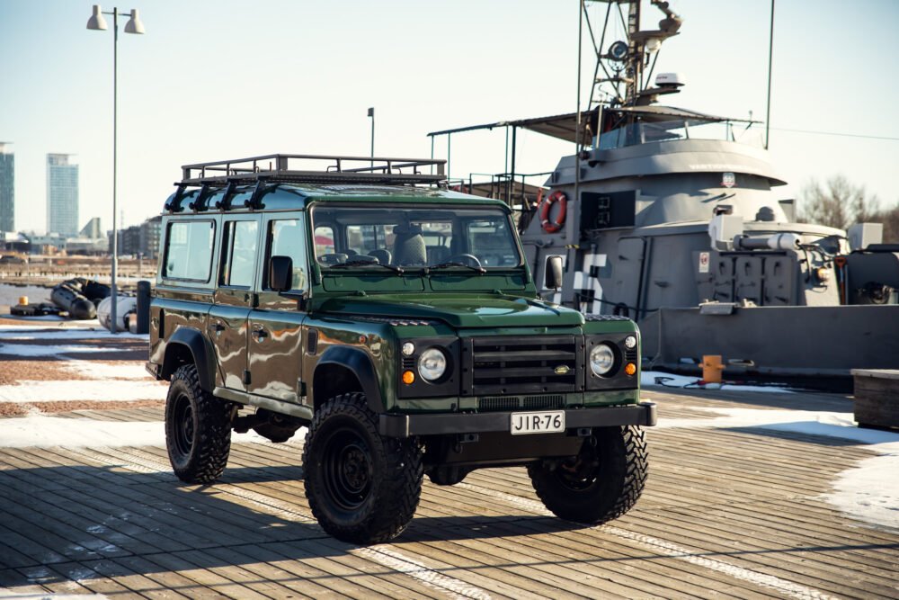 Green SUV near naval vessel in sunny dockyard.